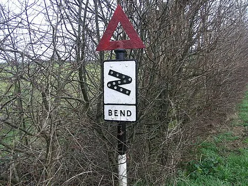 A weathered red triangular warning sign with a bend pictogram, mounted on a post beside a hedgerow on a rural English road.