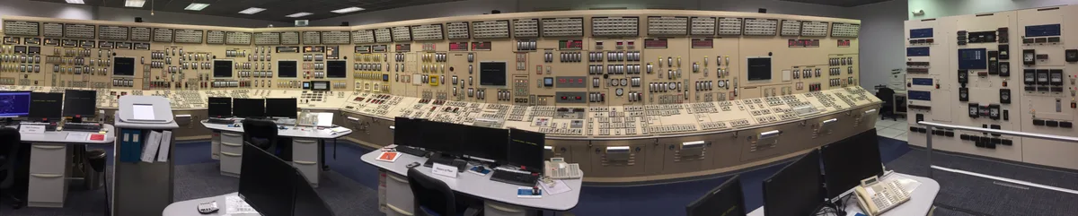 Panoramic view of a nuclear power plant control room: a long curved wall of near-identical beige panels covered in switches, gauges and indicator lights, with operator desks in front.