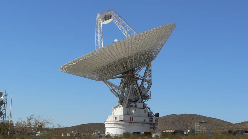 A large white dish antenna at NASA's Goldstone Deep Space Communications Complex, angled skyward against a clear blue sky.