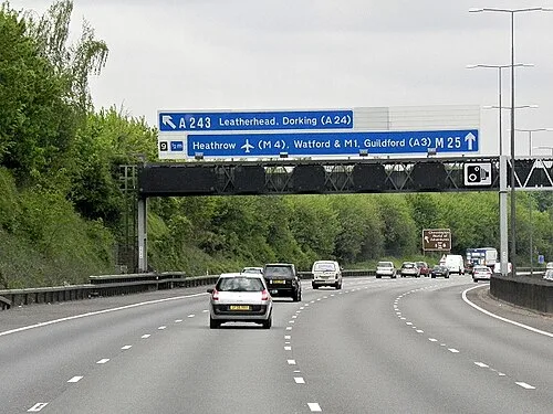 A gantry sign for the M25 and A243 seen from a moving car well before the Leatherhead junction, with a smaller advisory sign visible further down the carriageway.