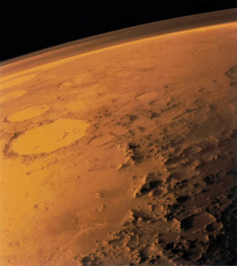 The curved limb of Mars seen from orbit, its thin atmosphere visible as a pale band against black space above cratered terrain.