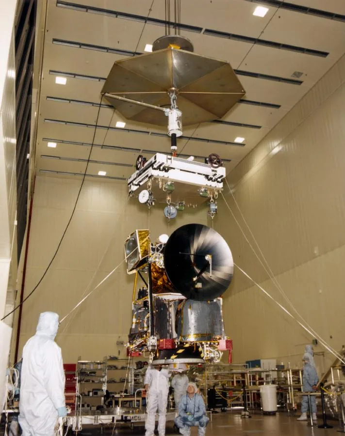 The Mars Climate Orbiter suspended by cables in a Lockheed Martin cleanroom, with technicians in white protective suits working below it.