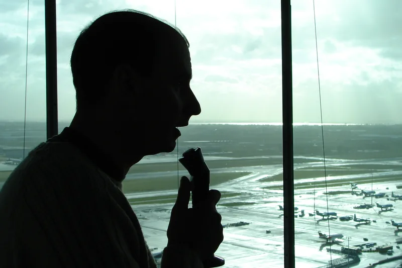 Silhouette of an air traffic controller speaking into a microphone at Amsterdam Schiphol Airport's control tower, with the airfield visible through the glass behind him.