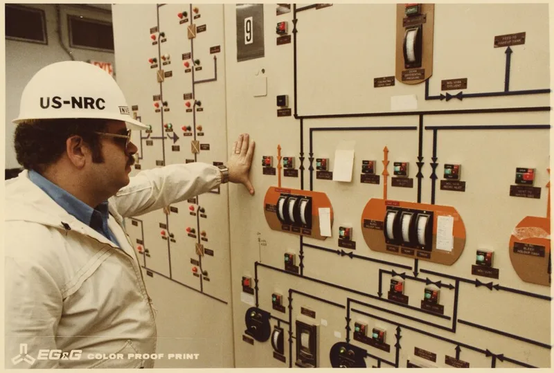 An NRC inspector in a hard hat reads a mimic panel of switches, indicator lights and process-flow lines on a nuclear control-room wall, hand resting on the panel.